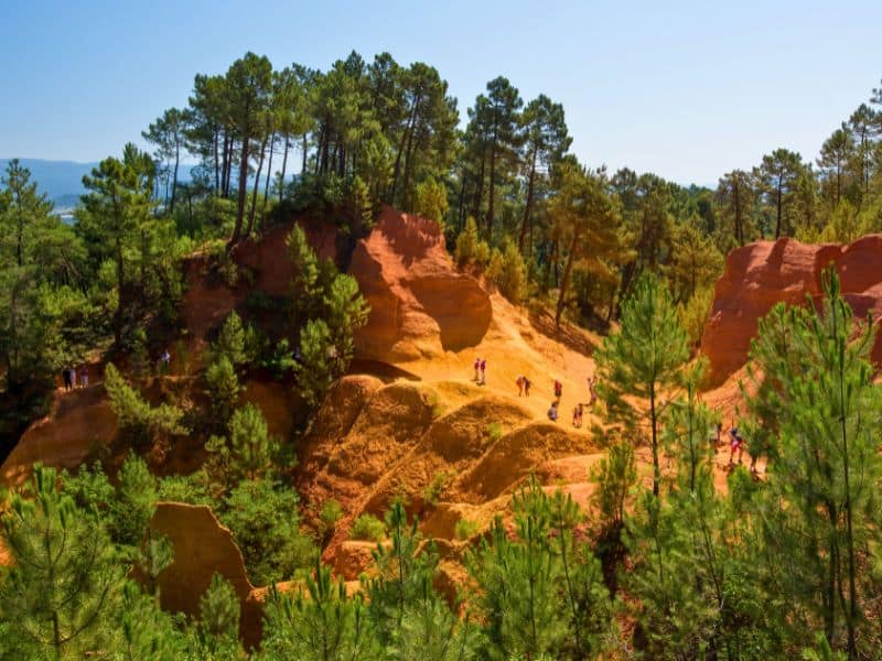 Paysage ocre du Luberon avec des falaises orangées, des pins et des visiteurs marchant sur les sentiers.