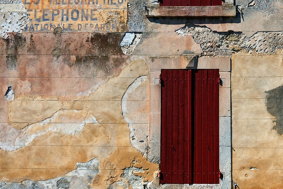Façade en pierre patinée de Ménerbes avec volets en bois rouge et anciennes inscriptions effacées.